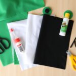 Materiały potrzebne do wykonania stroju żaby rozłożone na stole, w tym filc w kolorze zielonym, białym i czarnym, nożyczki, klej, drucik florystyczny i opaska